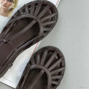 Puede incluir: Un par de sandalias de cuero marr&oacute;n con un dise&ntilde;o de tiras. Las sandalias tienen una suela plana y una puntera redondeada. Las tiras crean un patr&oacute;n de rayos de sol en la parte superior del pie. Las sandalias se muestran sobre una superficie blanca.