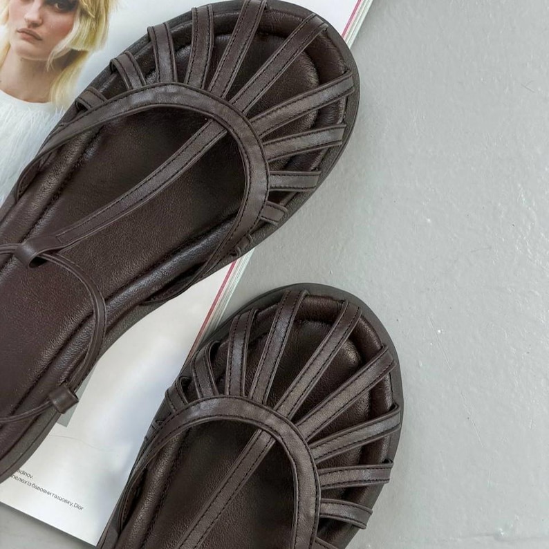 Puede incluir: Un par de sandalias de cuero marr&oacute;n con un dise&ntilde;o de tiras. Las sandalias tienen una suela plana y una puntera redondeada. Las tiras crean un patr&oacute;n de rayos de sol en la parte superior del pie. Las sandalias se muestran sobre una superficie blanca.