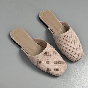 May include: A pair of light beige slip-on mules featuring a square toe design and a perforated upper surface. The interior footbed is a matching beige tone, and the shoes have a flat sole construction, providing a simple and clean aesthetic.