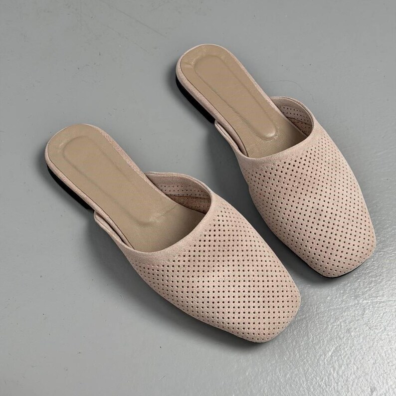 May include: A pair of light beige slip-on mules featuring a square toe design and a perforated upper surface. The interior footbed is a matching beige tone, and the shoes have a flat sole construction, providing a simple and clean aesthetic.