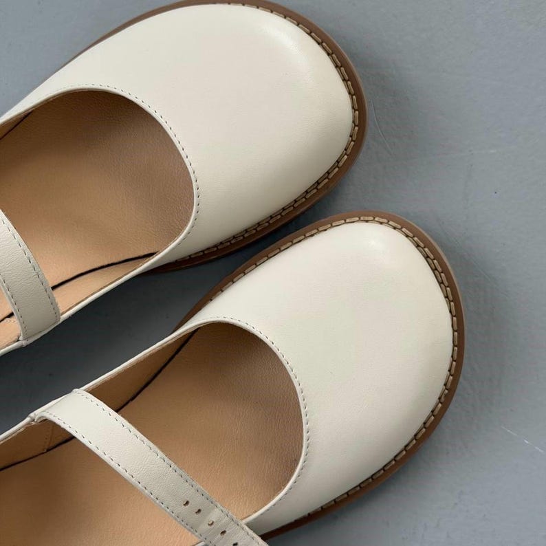 May include: Close-up of two white leather Mary Jane shoes. The shoes have a rounded toe, a strap across the top, and a brown sole. The stitching around the sole is visible. The shoes are on a grey surface.