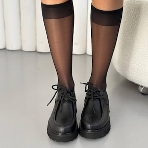 May include: Black leather shoes with thick soles and lace-up closures. The shoes are paired with sheer black knee-high stockings. The footwear has a classic design with a modern, chunky sole.
