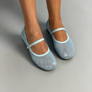 May include: A pair of light blue Mary Jane flats featuring a mesh upper construction with a fine grid pattern. Each shoe includes a thin, matching light blue strap across the instep with a small metal buckle closure and a rounded toe design.