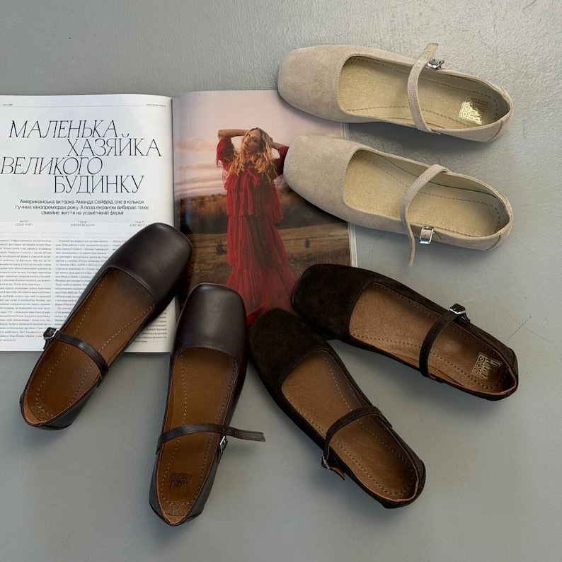 May include: An assortment of women's flats in various colors, including beige, brown, and black. The shoes have a strap across the top and are displayed with an open magazine. The text on the magazine is in a foreign language.