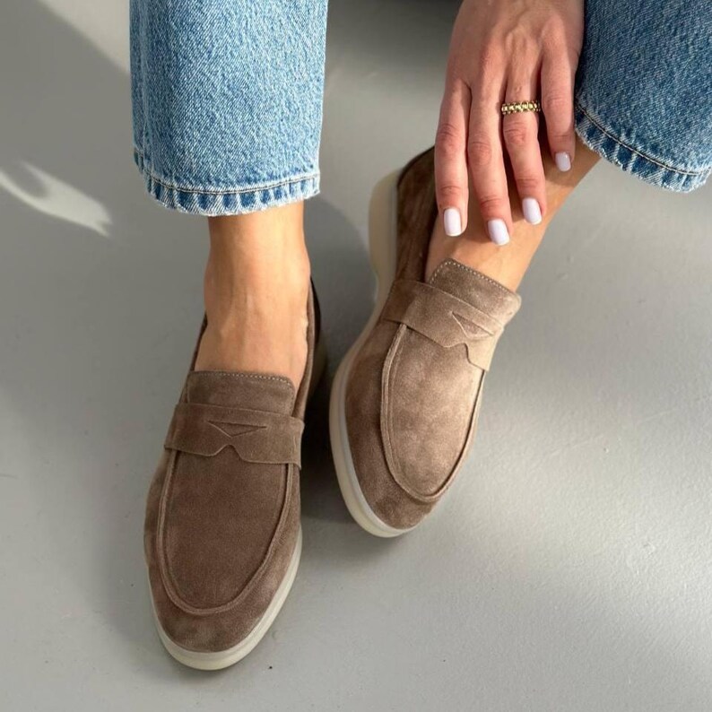 May include: A pair of brown suede loafers with a light-colored sole. The shoes have a classic design with a strap across the top. The image shows the shoes from a low angle, highlighting their shape and texture.
