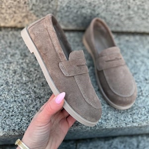 May include: A pair of taupe suede loafers with a slip-on design. The shoes have a rounded toe, a strap across the top, and a light-colored sole. The shoes are displayed on a stone surface.