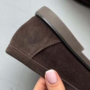 May include: A dark brown suede shoe featuring a textured finish and visible tonal stitching along the heel and side panels. The shoe has a flat, dark brown rubber sole with a clean edge, showcasing the material quality and construction details.