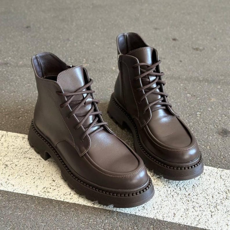 May include: A pair of brown leather ankle boots with a lace-up design and a chunky, ridged sole. The boots have a rounded toe and a zipper on the side. The boots are approximately 6 inches in height.