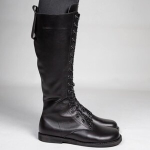 May include: A tall black leather boot featuring a full-length front lace-up closure with matching black eyelets and laces. The boot has a rounded toe, a flat sole, and a vertical pull tab at the top rear of the shaft, extending to just below the knee.
