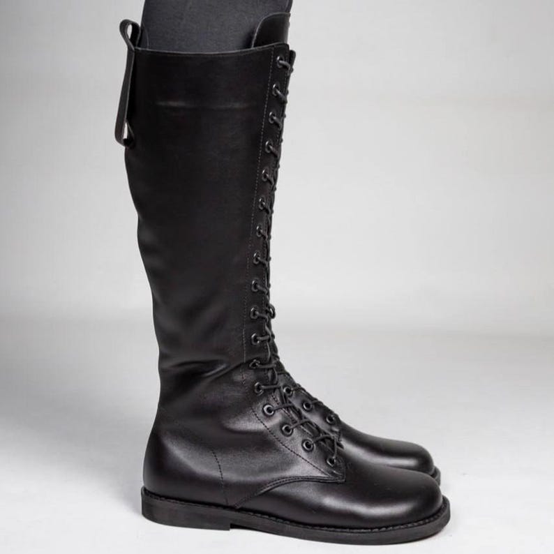 May include: A tall black leather boot featuring a full-length front lace-up closure with matching black eyelets and laces. The boot has a rounded toe, a flat sole, and a vertical pull tab at the top rear of the shaft, extending to just below the knee.