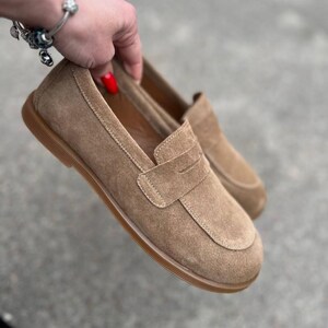 May include: A pair of tan suede loafers with a rounded toe and a penny strap detail. The shoes have a brown rubber sole and are held up by a hand. The shoes are a neutral color and are suitable for casual wear.
