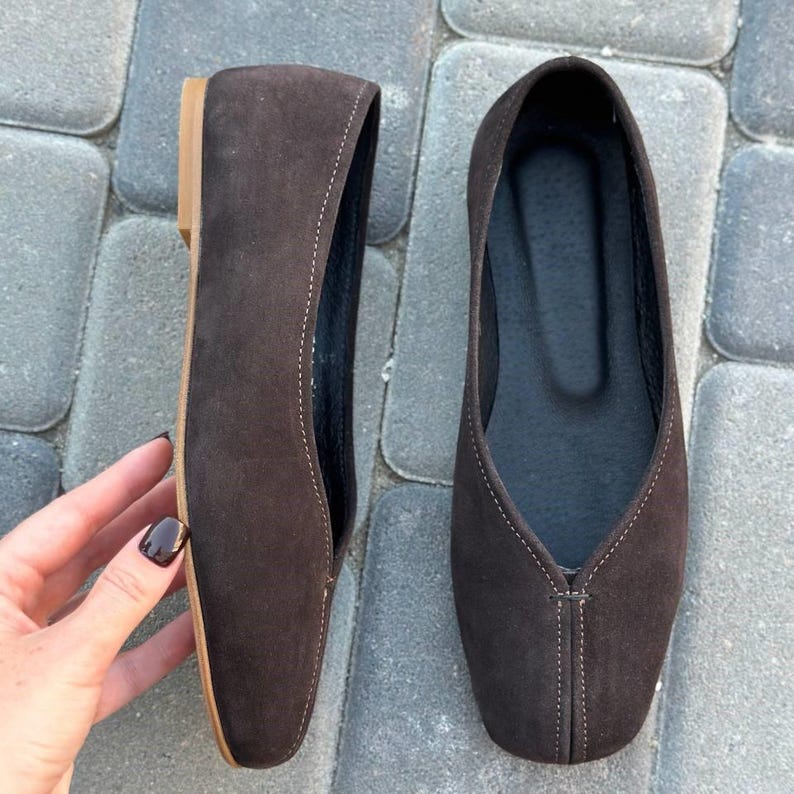 Pode incluir: Duas sapatilhas de camur&ccedil;a castanhas com bico fino e salto baixo. Os sapatos t&ecirc;m detalhes de costura e interior castanho escuro. Uma m&atilde;o segura um sapato, mostrando a forma e o design.