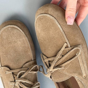 May include: Tan suede moccasin-style shoes with laces. The shoes have a rounded toe and a stitched design around the top. The laces are tied in a bow. The shoes are a neutral color and appear to be unworn.
