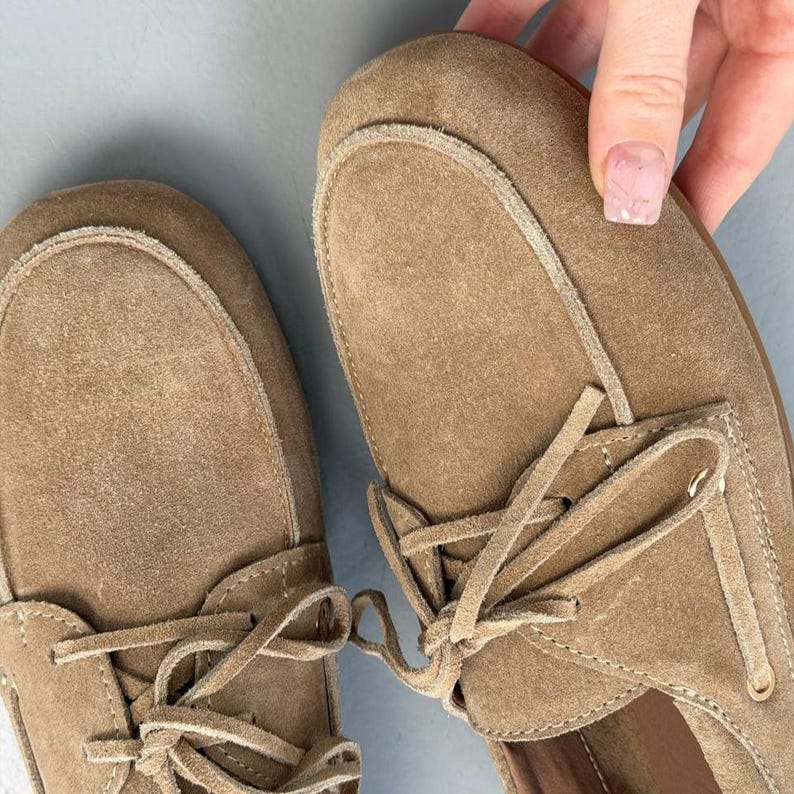 May include: Tan suede moccasin-style shoes with laces. The shoes have a rounded toe and a stitched design around the top. The laces are tied in a bow. The shoes are a neutral color and appear to be unworn.