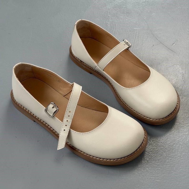 May include: A pair of cream-coloured Mary Jane shoes with a strap and buckle fastening. The shoes have a rounded toe and a brown sole. The shoes are made of a smooth material and appear to be leather.