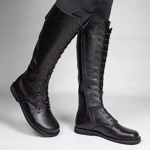 May include: A pair of black knee-high boots featuring a full-length front lace-up closure with black eyelets and laces. The boots have a rounded toe, a flat sole, and a vertical side zip on the inner shaft for closure, extending to the knee.