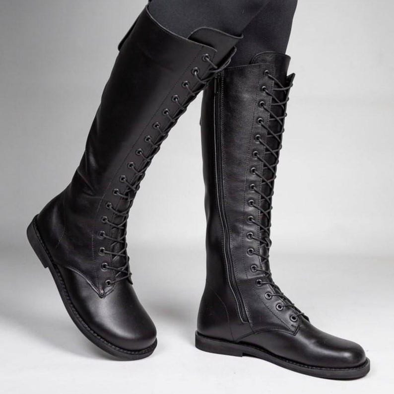 May include: A pair of black knee-high boots featuring a full-length front lace-up closure with black eyelets and laces. The boots have a rounded toe, a flat sole, and a vertical side zip on the inner shaft for closure, extending to the knee.
