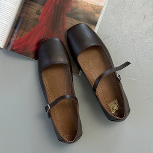 May include: A pair of dark brown leather Mary Jane flats with a square toe and a strap with a buckle closure. The shoes have a light brown interior and are set on a gray surface.