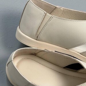 May include: Close-up of two off-white leather shoes. The shoes have a rounded toe, a low profile sole, and a small elastic panel on the side. The interior of the shoe is a lighter shade of off-white.