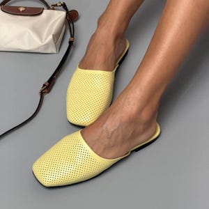 May include: A pair of light yellow slip-on mules featuring a perforated texture across the upper surface, a square toe design, and a thin black sole, accompanied by a small white and brown handbag with a long adjustable shoulder strap.