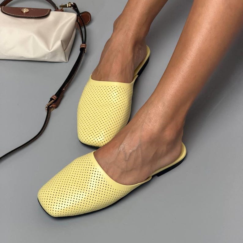 May include: A pair of light yellow slip-on mules featuring a perforated texture across the upper surface, a square toe design, and a thin black sole, accompanied by a small white and brown handbag with a long adjustable shoulder strap.