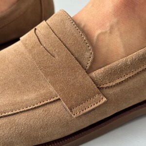 May include: A tan suede penny loafer featuring a classic strap across the vamp with a horizontal slit. The shoe has visible tonal stitching along the edges and a dark brown sole. The texture of the suede is matte and uniform in color throughout.