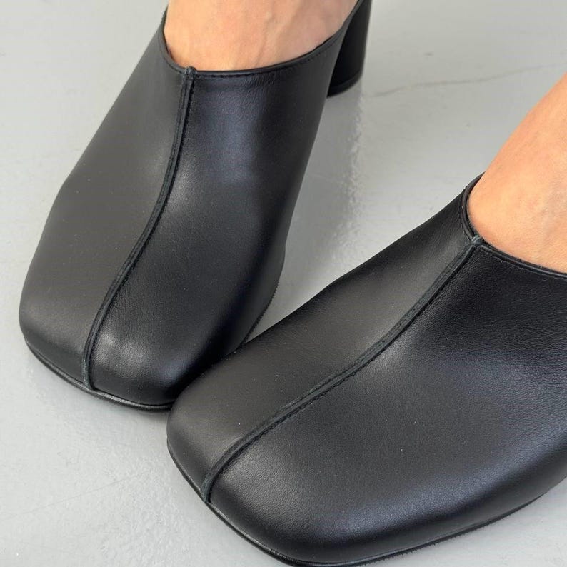 May include: Close-up of black leather mules with a square toe and block heel. The shoes have a minimalist design with a seam detail running down the center. The background is a light gray surface.