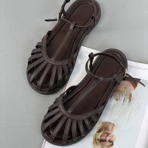 Puede incluir: Un par de sandalias de cuero marr&oacute;n con dise&ntilde;o de tira en T y m&uacute;ltiples correas en el pie. Las sandalias tienen suela plana y correas ajustables en el tobillo con hebillas doradas. Las sandalias se muestran sobre una superficie gris claro.