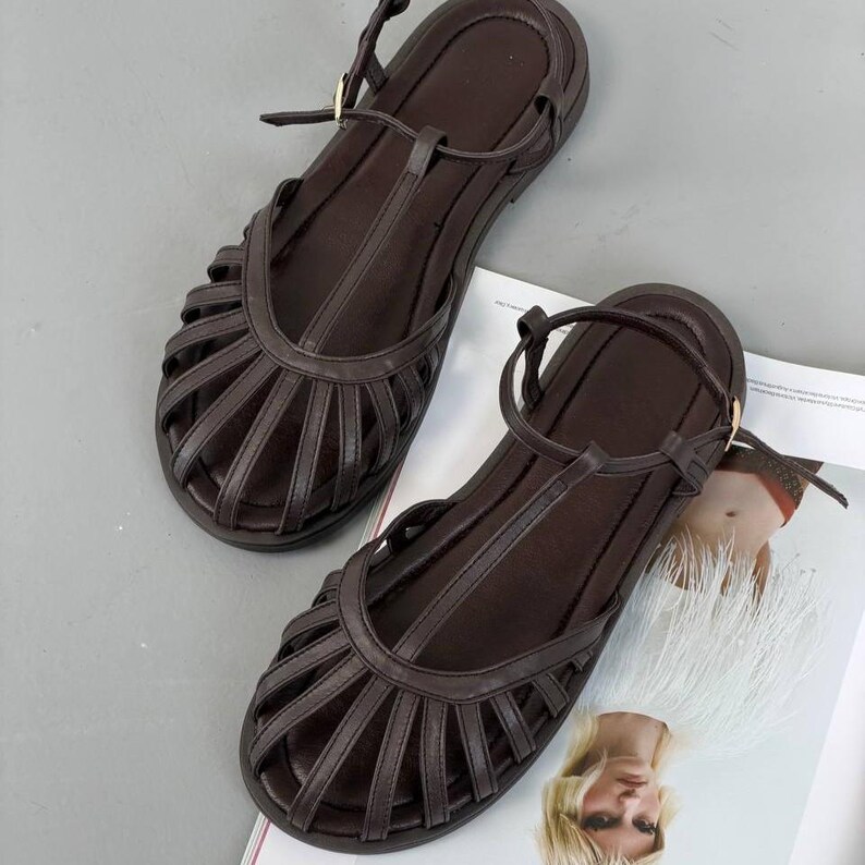Puede incluir: Un par de sandalias de cuero marr&oacute;n con dise&ntilde;o de tira en T y m&uacute;ltiples correas en el pie. Las sandalias tienen suela plana y correas ajustables en el tobillo con hebillas doradas. Las sandalias se muestran sobre una superficie gris claro.