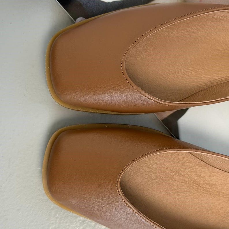 May include: Close-up of two brown leather flats with a square toe design. The shoes have a smooth, polished finish and a light-colored sole. The interior of the shoes is a lighter shade of brown.