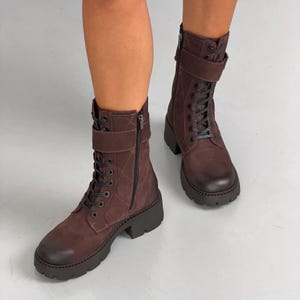 Könnte beinhalten: Dunkelbraune wadenhohe Stiefel mit gestepptem Rautenmuster am Schaft, schwarzer Schnürung und seitlichem Reißverschluss. Die Stiefel verfügen über einen Riemen mit Schnalle am oberen Knöchel sowie eine dicke, profilierte Gummisohle mit Blockabsatz.