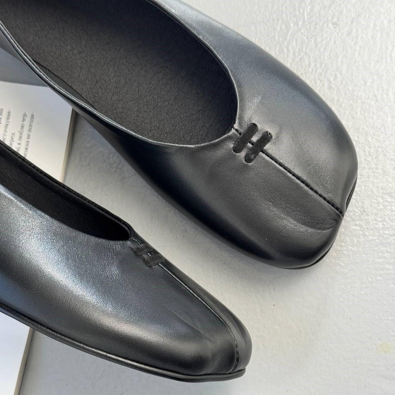 Op de afbeelding: Close-up van twee zwarte leren ballerina's. De ballerina's hebben een vierkante neus en een klein decoratief stikseldetail op de wreef. De schoenen staan op een lichtgekleurd oppervlak.
