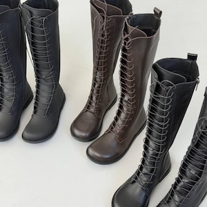 May include: A collection of knee-high boots featuring a lace-up front design with matching laces, rounded toes, and side zips. The boots are available in solid black and dark brown colours, showcasing a smooth finish and a flat sole construction.