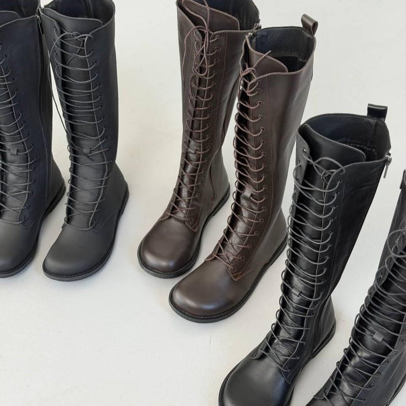 May include: A collection of knee-high boots featuring a lace-up front design with matching laces, rounded toes, and side zips. The boots are available in solid black and dark brown colours, showcasing a smooth finish and a flat sole construction.