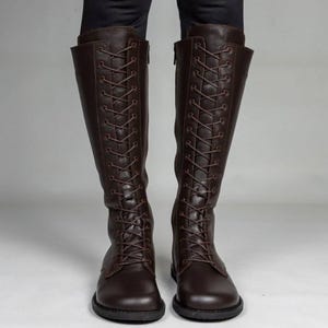 May include: A pair of dark brown knee-high boots featuring a full-length front lace-up design with matching brown laces, rounded toes, and a side zip closure. The boots have a smooth finish and a flat sole, extending up to the lower calf area.