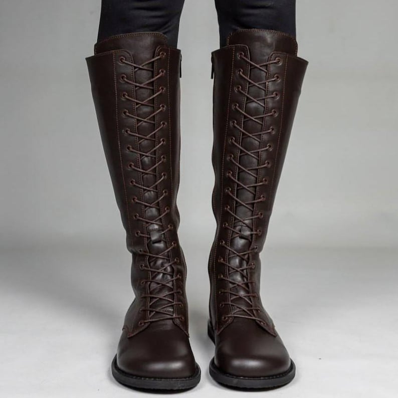 May include: A pair of dark brown knee-high boots featuring a full-length front lace-up design with matching brown laces, rounded toes, and a side zip closure. The boots have a smooth finish and a flat sole, extending up to the lower calf area.
