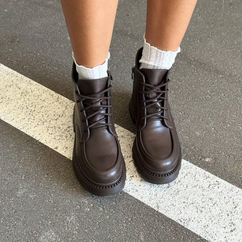 May include: Brown leather ankle boots with a lace-up design and a chunky sole. The boots have a rounded toe and a zipper on the side. White socks peek out from the top of the boots. The boots are on a gray and white striped surface.