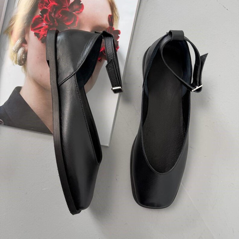 May include: A pair of black leather flats with ankle straps. The shoes have a square toe design and a low heel. The straps are secured with small silver buckles. The flats are displayed on a light gray surface.