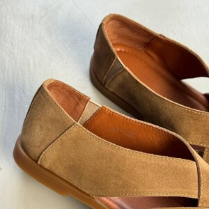 May include: Close-up of two tan suede flats. The shoes have a criss-cross design over the foot and a brown sole. The interior of the shoe is a lighter shade of brown. The shoes are on a white surface.