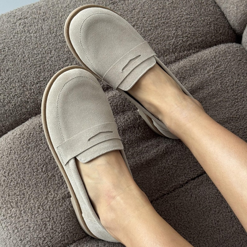 May include: A pair of light beige suede loafers featuring a classic penny slot strap across the vamp, a rounded toe design, and a thin brown sole. The shoes have a soft, matte texture and a slip-on construction with a clean, simple silhouette.
