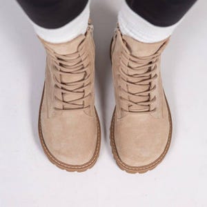 Könnte beinhalten: Ein Paar beigefarbene Schnürstiefel aus Wildleder mit abgerundeter Zehenpartie, passenden beigen Schnürsenkeln und einer dicken, strukturierten Gummisohle mit sichtbaren Nähten entlang des Rahmens. Die Stiefel reichen über den Knöchel.