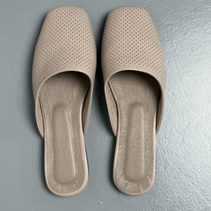 May include: A pair of beige slip-on mules featuring a square toe design and a perforated upper surface. The interior footbed is smooth and matches the exterior color, providing a clean and uniform appearance for these flat, open-back shoes.
