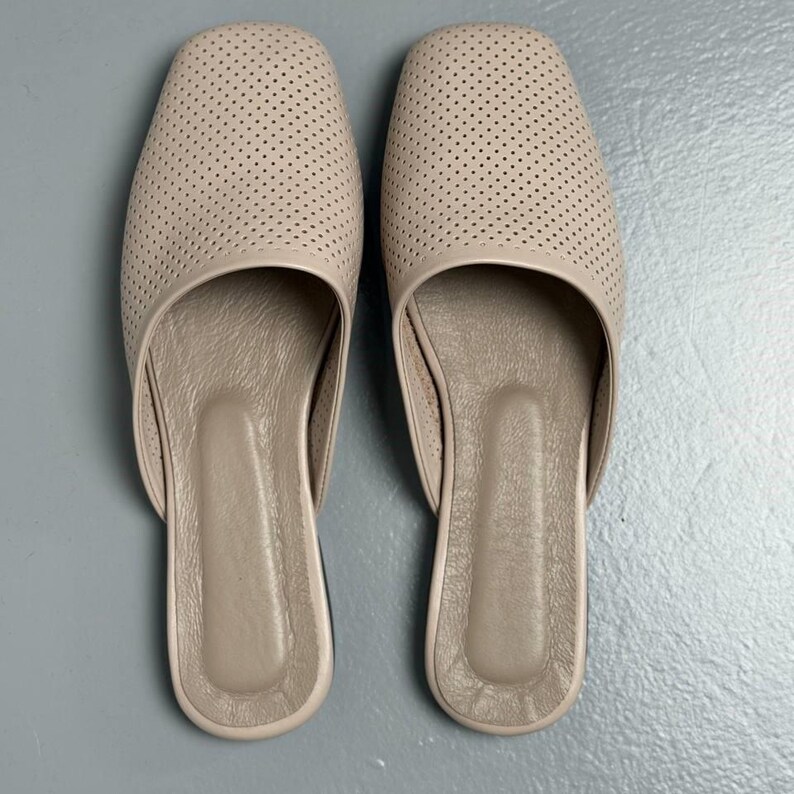 May include: A pair of beige slip-on mules featuring a square toe design and a perforated upper surface. The interior footbed is smooth and matches the exterior color, providing a clean and uniform appearance for these flat, open-back shoes.