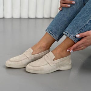 May include: A pair of beige suede loafers featuring a classic penny slot strap across the vamp, a rounded toe, and a flat, tonal rubber sole. The shoes are crafted from a soft, matte material with visible stitching along the moccasin-style toe seam.