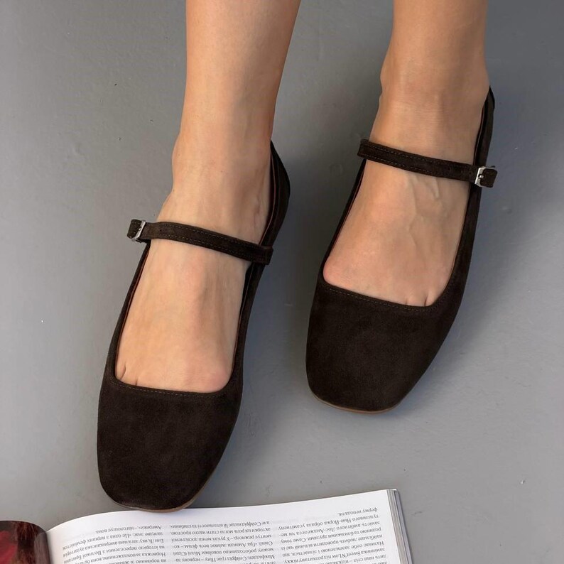 May include: Dark brown suede Mary Jane flats with a square toe and a buckle strap. The shoes are displayed on a gray surface, showcasing their simple, elegant design. These flats offer a classic style.