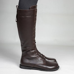May include: A knee-high dark brown boot featuring a full-length lace-up front with matching brown laces and metal eyelets. The boot has a smooth finish, a rounded toe, a flat sole, and a vertical pull tab at the top rear of the shaft.