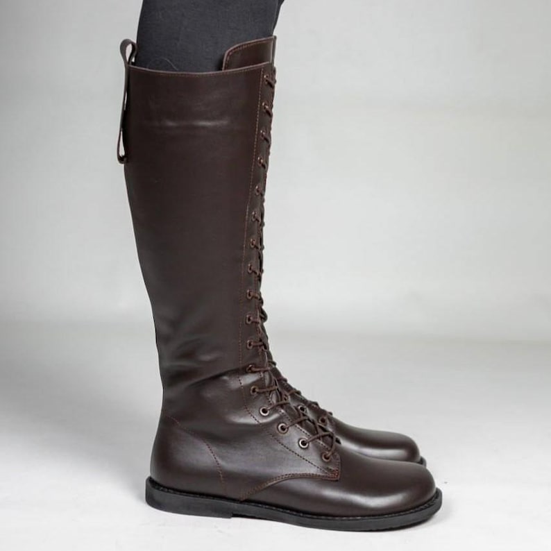May include: A knee-high dark brown boot featuring a full-length lace-up front with matching brown laces and metal eyelets. The boot has a smooth finish, a rounded toe, a flat sole, and a vertical pull tab at the top rear of the shaft.