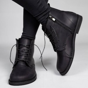 May include: A pair of matte black leather ankle boots featuring a rounded toe, thin black laces, and a silver-toned metal side zip. The boots have a low heel and a smooth, clean finish, extending slightly above the ankle with a structured design.