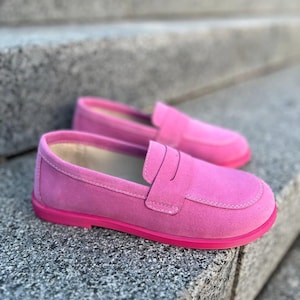 May include: A pair of bright pink suede loafers with a rounded toe and a strap across the top. The shoes have a matching pink sole and are set against a gray stone background.