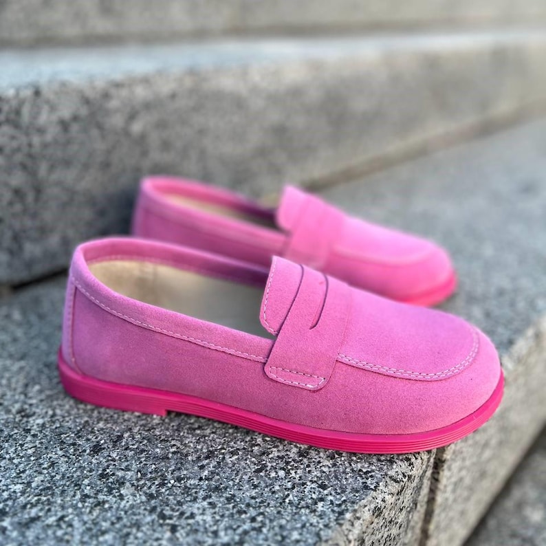 May include: A pair of bright pink suede loafers with a rounded toe and a strap across the top. The shoes have a matching pink sole and are set against a gray stone background.